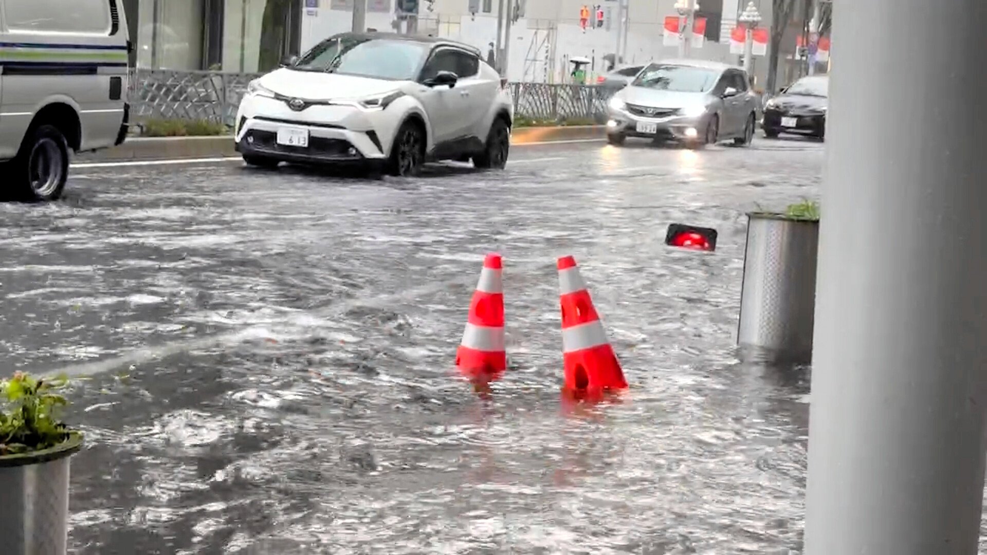 A car driving through a heavily flooded street, sending waves of water washing over the pavement